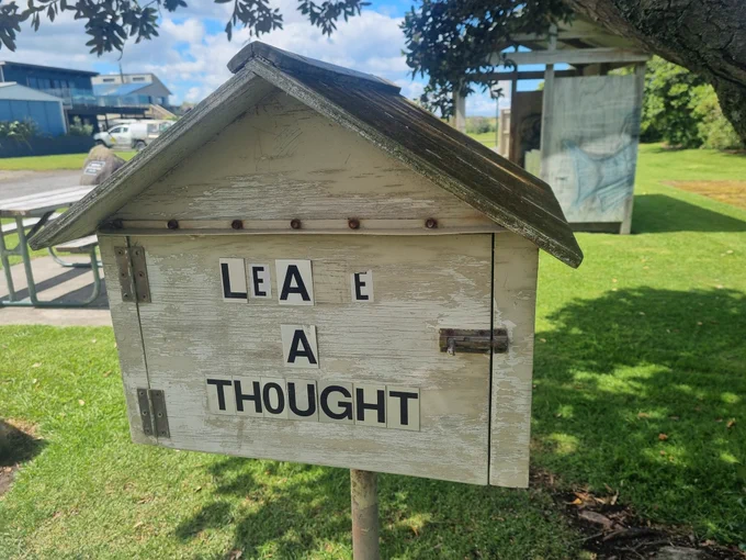 This photograph depicts a small birdhouse-style wooden box on a bright sunny day with a bluebird sky in the background. Someone has labeled the hinged front door with the phrase "leave a thought" though the V sticker has peeled away. Weathered and well-loved, it has seen some use.
