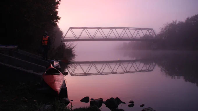 Ein Bild des Wesel-Datteln-Kanals bei Morgengrauen. Eine Person steht neben einem roten Kayak am Ufer. Im Hintergrund eine Brücke.