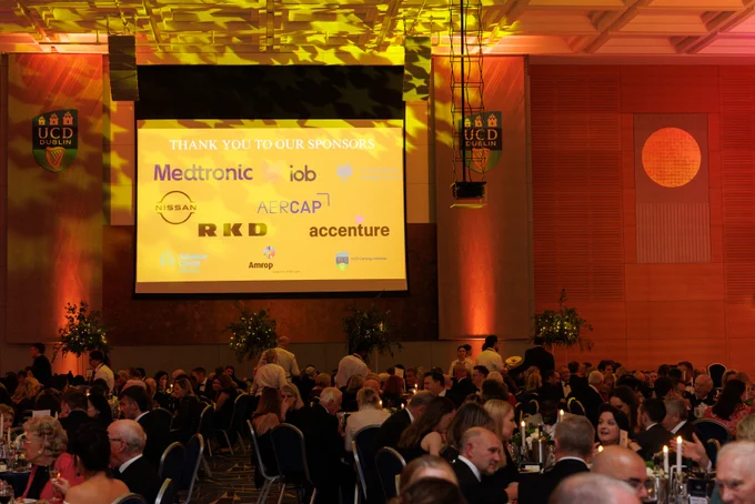 A projector screen displaying the logos of UCD Alumni Awards sponsors, with the text 'Thank you to our sponsors' at the top. The room is filled with attendees seated at round tables during the awards event.