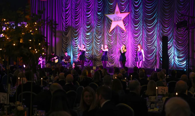 A group of dancers performing on stage at the UCD Alumni Awards event, with vibrant purple and blue lighting against a draped curtain backdrop. Musicians are seated in the background, and a large star-shaped sign with 'UCD Alumni Awards' is prominently displayed.
