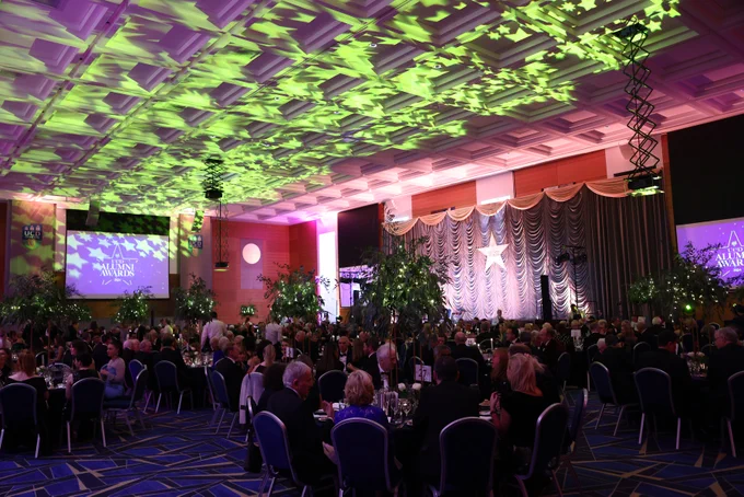 A wide view of the 2024 UCD Alumni Awards event, showing a grand ballroom with attendees seated at round tables under soft green and purple lighting.