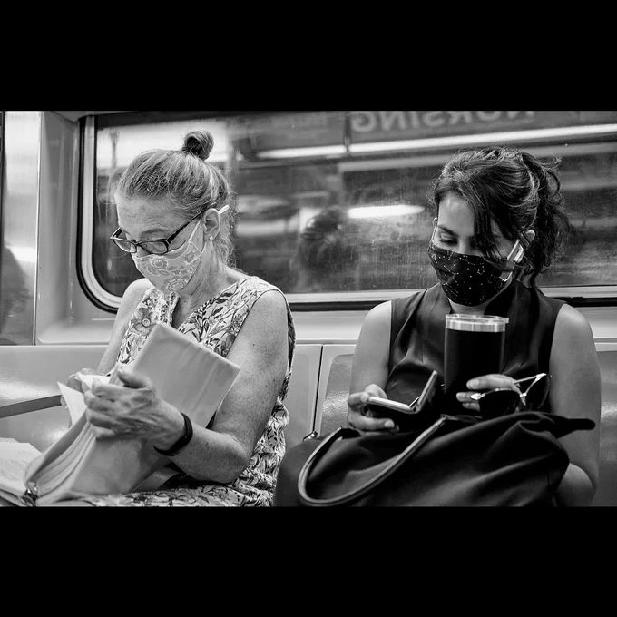 Two women seated side by side on a subway car, one looking at a book and the other looking at her phone.