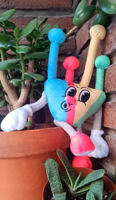 Peluche de Biznaga Fest 2004, con la forma de la mascota de la edición y los colores de Google. Está entre dos macetas llenas de plantas , con fondo de ladrillos