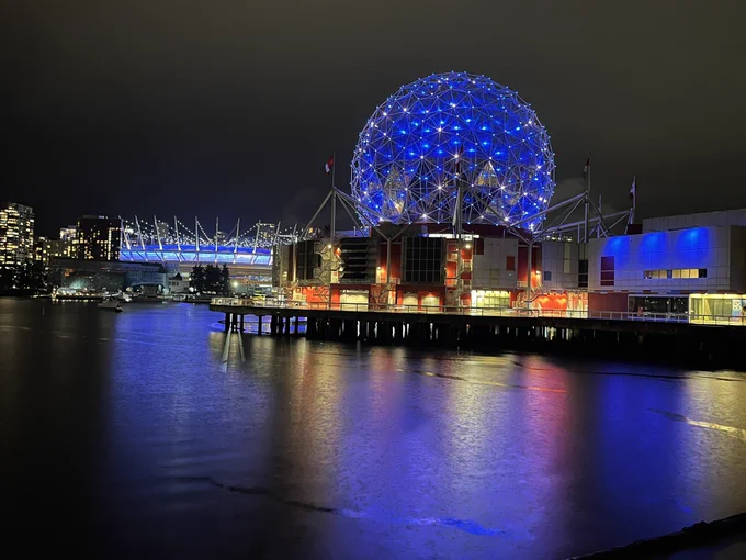 False Creek (body of water) in foreground with coloured lights reflecting on surface, geodesic dome (Science World) lit in blue in background, with stadium lit in blue to the left