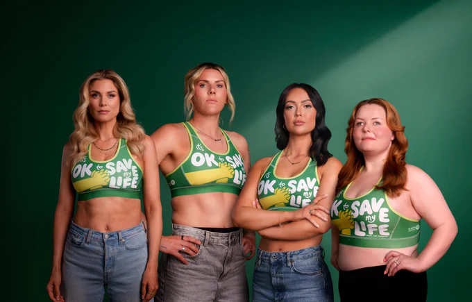 Photograph. Four women stand in front of a dark green backdrop. From left to right, Ashley James, Millie Bright, Sharon Gaffka, Lucy Edwards. They look strong and confident, with a determined look. They are all wearing jeans and green sports bras that read "It's okay to save my life". The bras have a yellow hand illustration, representing how someone would perform CPR