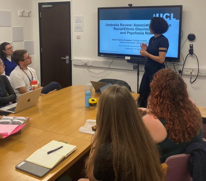 India Francis-Crossley from UCL presents her research at today's session of the Race and Psychiatry Journal Club at the University of Oxford's Department of Psychiatry. She is standing next to a screen listened to by people sitting around a table.
