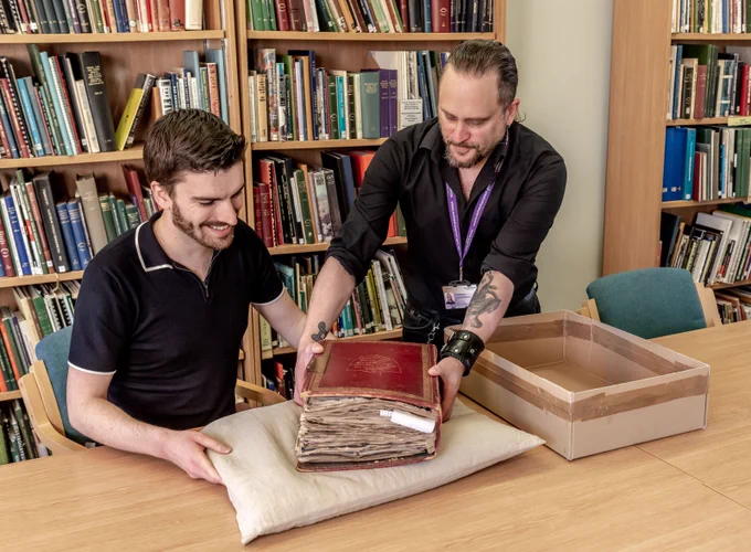 One person delicately places a large book on to cushion laid out on a table. Another person is sat at the table ready to read the book. There are shelves with more books behind them.