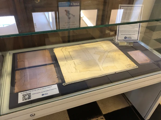 A photograph of an exhibition cabinet in the foyer of York Explore. Inside there is a large plan held down with weights. There are also photographs of the plan before it underwent conservation work and text to accompany it. There is also a QR code to donate.