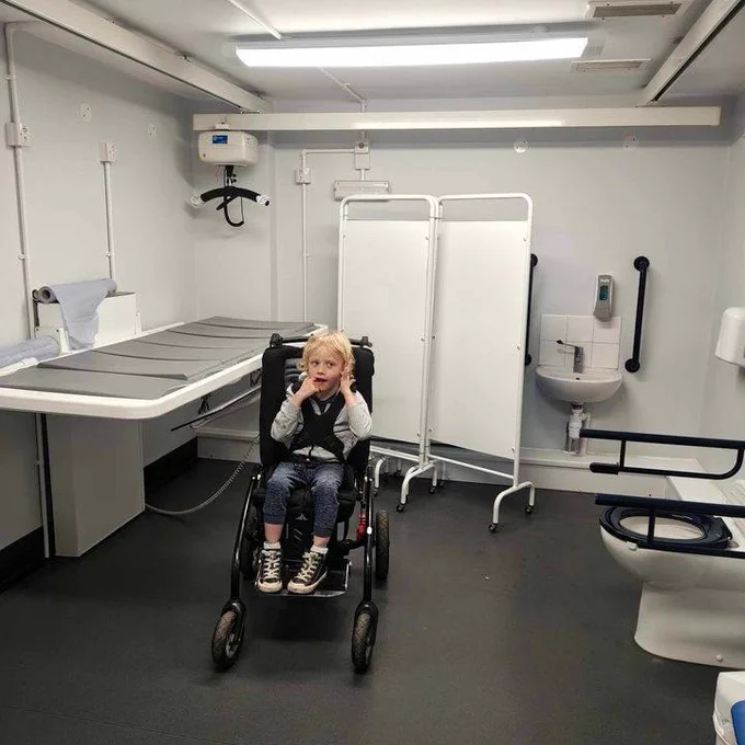 Ryder has blonde hair and is sitting in his specialist chair inside the Changing Places toilet. It has a changing bench, toilet, hoist, sink and privacy screen on display. The floor is grey and the walls are white.