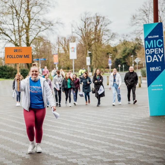 A female MIC student leading a group on a campus tour at the MIC Open Days. She is wearing a blue MIC branded t-shirt, red leggings and a white jacket. She is holding an orange sign that says 'Follow Me'. There is also a blue and navy sign wrapped around a pole that says 'MIC Open Day'. 
