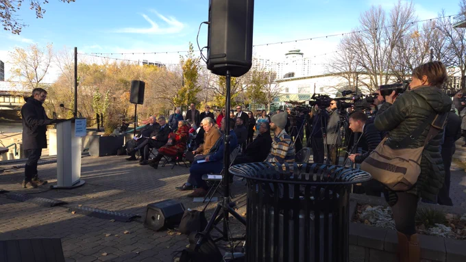 Man, on the left, talking into a microphone with people, with and without cameras, standing and sitting down in front of him. 