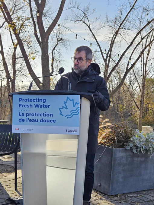 man outside, with plants trees behind him, talking into a microphone. Text, sign on the podium: Protecting Fresh Water. La protection de l’eau douce