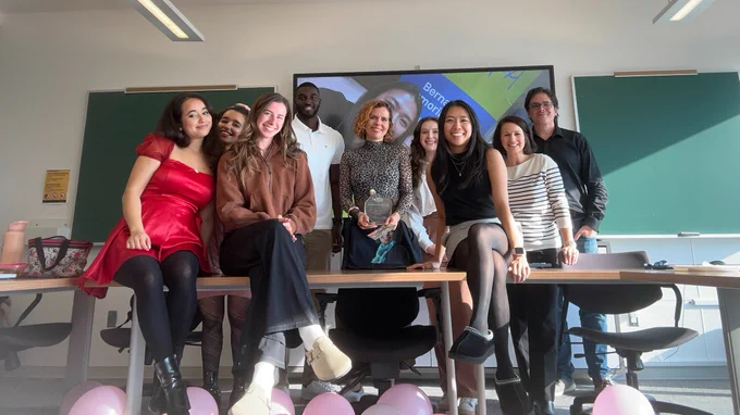 A group of people stand behind and sit on a table. A woman in the middle, Dr. Sophie Lebel, holds a glass plaque for the Bernard Fox Award. Happy people!!! There are also pink balloons on the floor.