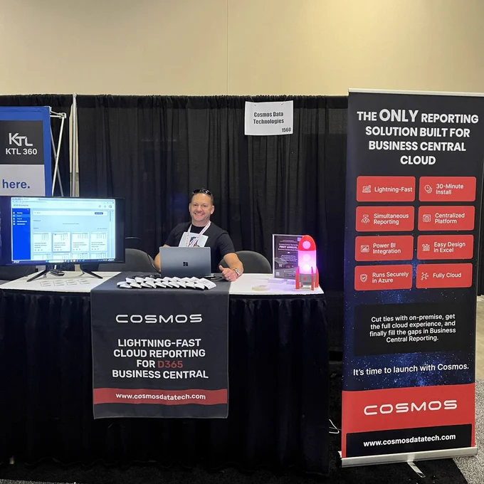 The Cosmos booth at Community Summit North America 2024 with Brian Petersen sitting behind the table. The booth displays signage promoting "Lightning-Fast Cloud Reporting for D365 Business Central," and features a banner with key benefits of their reporting solution. A small lit rocket model adds a space-themed touch to the setup.