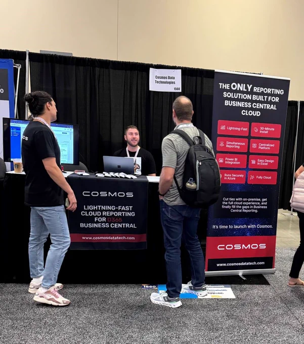 Anthony Bonaduce is behind the Cosmos booth, engaging with a visitor. Andrew Pierson is standing on the left.  The booth displays banners promoting Cosmos' cloud reporting solution for Business Central, with a small lit rocket model on the table.
