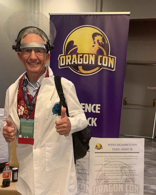 a man in a lab coat and safety visor is flashing thumps up at the camera while standing in front of a big sign for Dragon Con