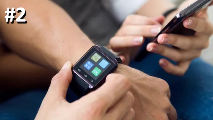 The image shows a close-up of a person's wrist wearing a smartwatch. The smartwatch screen displays four app icons: a clock, a calendar, a calculator, and a messaging app. The person is interacting with the smartwatch using their other hand. In the background, another person is holding a smartphone. The number "#2" is visible in the top left corner of the image.