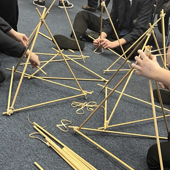 People sitting on a floor build structures using wooden poles
