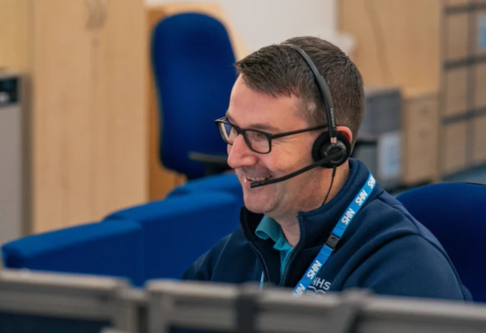 A smiling NHS 24 call handler sitting at their desk with a headset on.