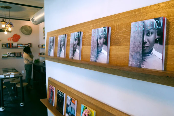 A photo of a display of Seen Issue 007, they are lined up on a wall shelf, beneath are copies of early editions of Seen. In the distance someone is shopping. 