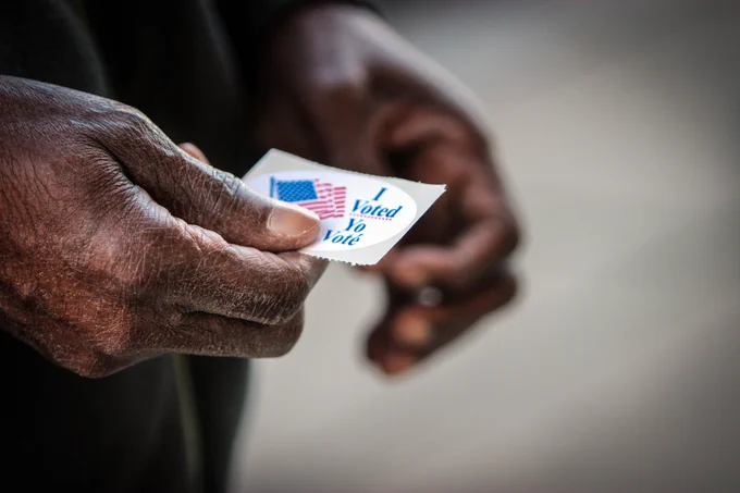 A person holds a small sticker that reads "I Voted" with an American flag design.