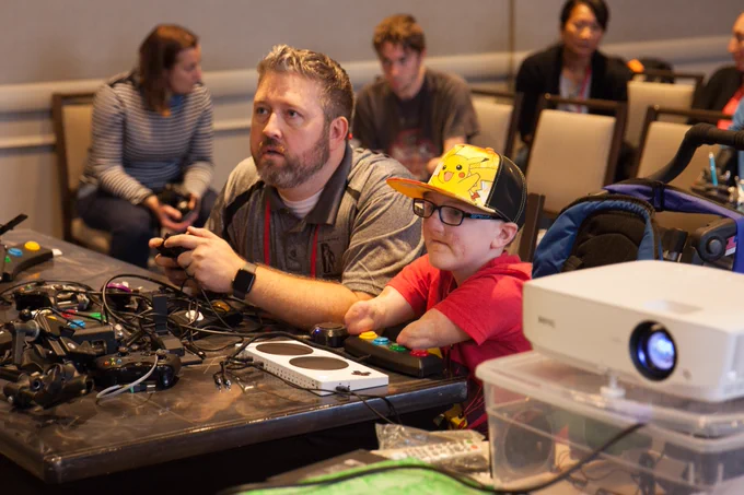 A man (me, Erik Johnson) and a young boy are seated at a table, both focused on playing video games. The boy, wearing a Pikachu-themed cap and glasses, uses the xbox adaptive controller and adaptive switches to play. The table in front of them is filled with various adaptive gaming devices and wires. In the background, several other people observe. 
