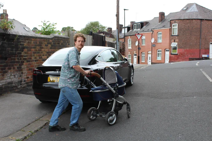 A man wearing a blue patterned shirt and jeans pushes a pram around a car. The car is parked on the pavement and blocking access. 