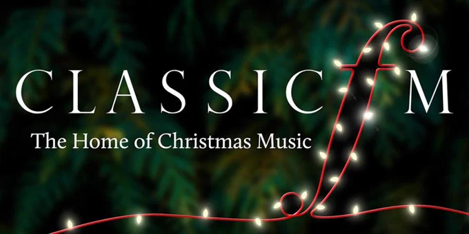 The image is a banner for Classic FM, featuring the text "Classic FM" with the tagline "The Home of Christmas Music." The design incorporates festive elements, with lights wrapped around the letter "f" in "FM" and a dark, green background resembling foliage, evoking the spirit of the holiday season.