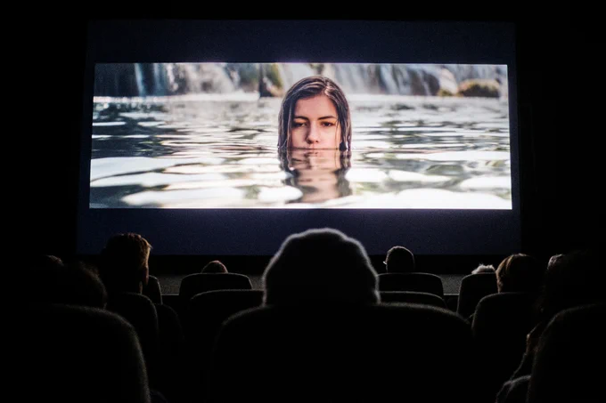 A photograph of the dark outline of an audience sitting in a cinema, with an image on the screen of a woman’s head emerging from a lake