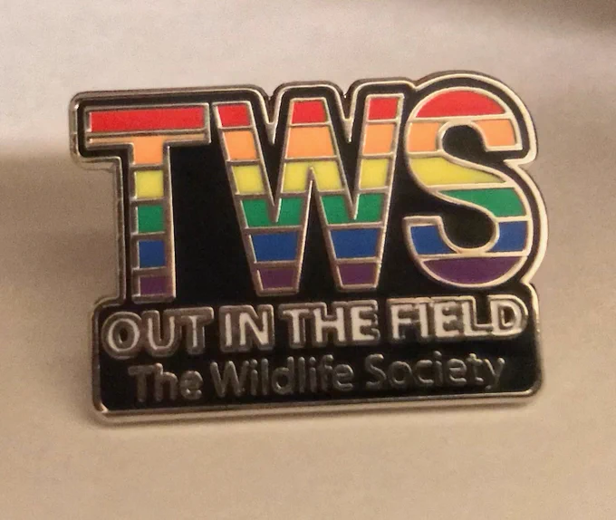 a small pin with the OiTF logo. The logo has “TWS” in capital letters and rainbow colors, with “OUT IN THE FIELD” below in white bold text and “The Wildlife Society” below that.