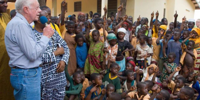 Former U.S. President Jimmy Carter speaking to a group of children and adults in a village.