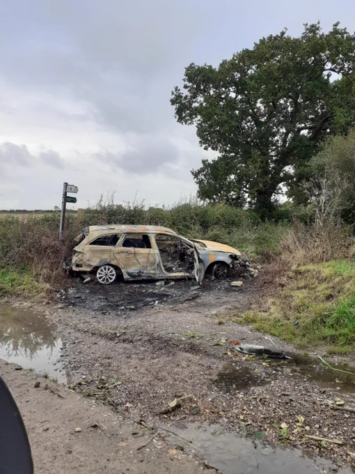 The image shows a severely burned-out car, left abandoned on a rural roadside. The vehicle's exterior is charred, and the front end seems particularly damaged. It is positioned near overgrown vegetation and a large tree, with a muddy patch in the foreground. There is also a small signpost in the background, which appears to mark a footpath or country road, though it is partially obscured. The scene conveys a desolate and somewhat neglected environment.

