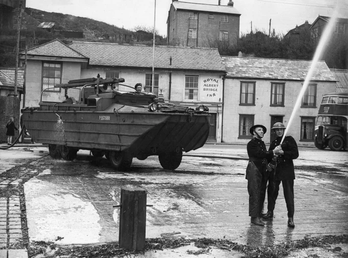 Two firefighters using a hose in front of a DUKW amphibious vehicle, which has been converted into a fire engine 