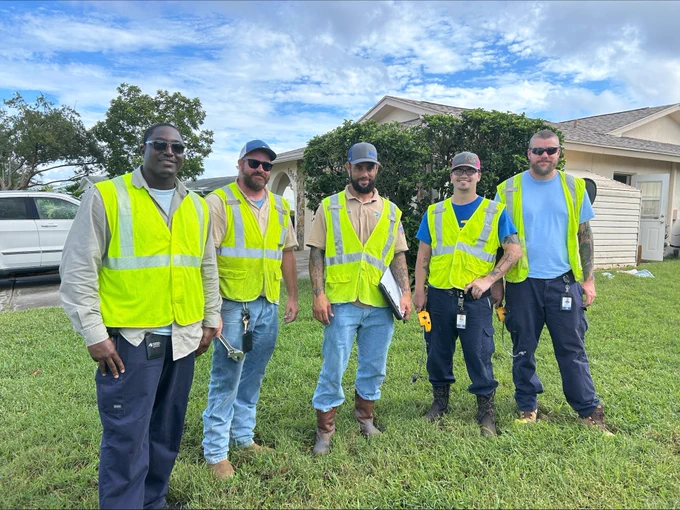 Five Peoples Gas workers in reflective safety vests stand in front of a residential house. They are equipped with various work tools.