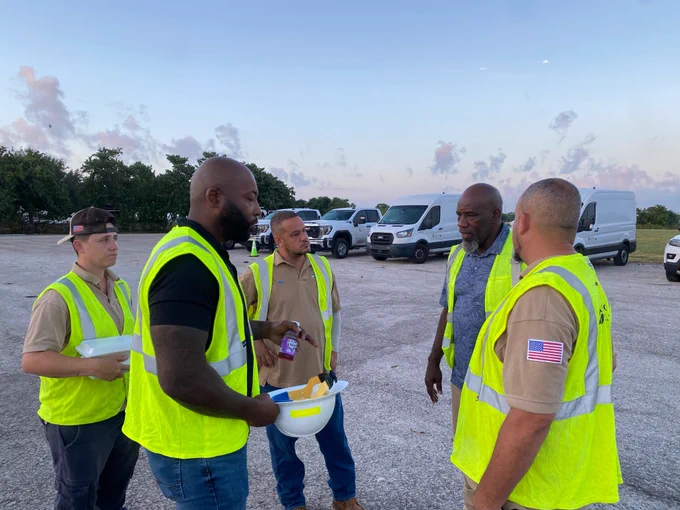 A group of Peoples Gas workers in safety vests engaging in a conversation in a parking area during dusk.