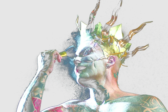 A dramatically lit photograph of the artist (a white man) against a black background, taken from a low angle, showing only his head and shoulders. He wears a gold metal crown, on which elastic cords are attached to hooks in his face, pulling his skin upwards. He is applying white powder to his face