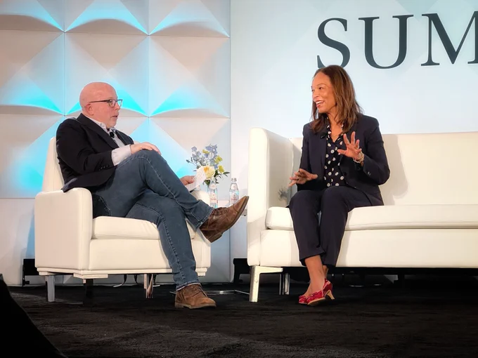 Two people engaged in a conversation on stage at a summit, with one seated on a white armchair and the other on a white sofa. The backdrop features a geometric design and the words "STAT SUMMIT."