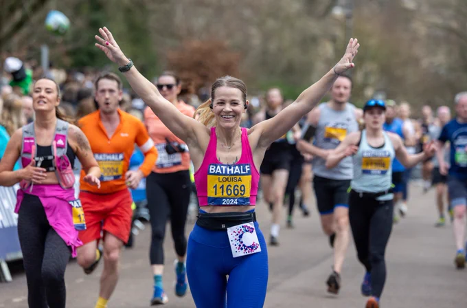 A photo of a smiling female finisher with her hands in the air on the home straight of the 2024 Bath Half.