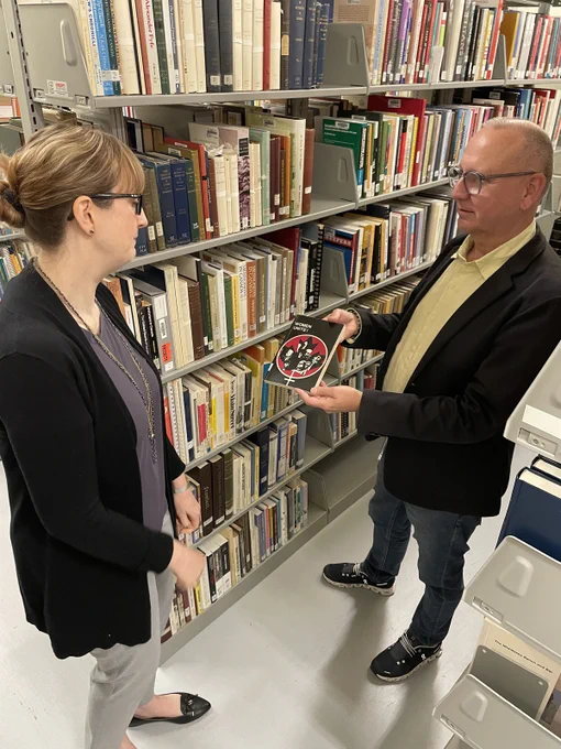 Two people standing in the stacks of a library looking at a book about women's liberation movements. 