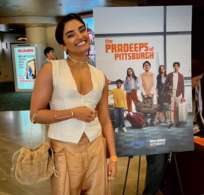 The photo shows Sahana Srinvasan wearing a white sleeveless top, beige pants and yellow hand bag standing next to a  promotional poster for “The Pradeeps of Pittsburgh” TV show. The poster shows a South Austin family with luggage. In the background, there is another poster for a movie called “Hellraisers”.Photo courtesy of Sahana Srinvasa.