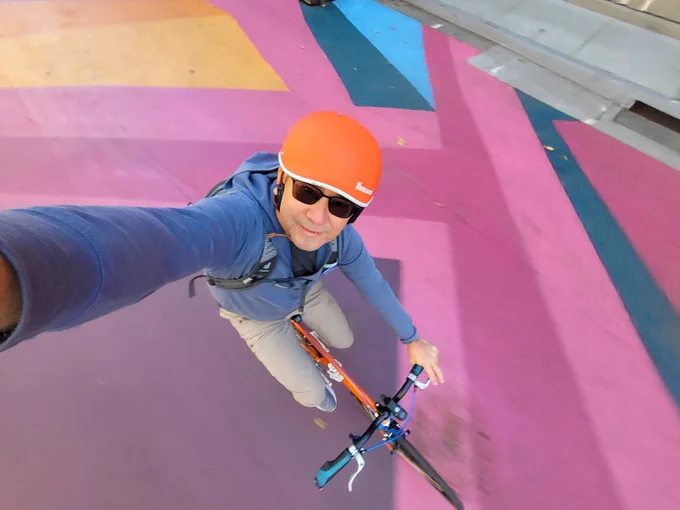 Wide angle selfie of Yours Truly on a bicycle. I'm wearing an orange bike helmet, a blue hoodie jacket, and khaki pants. The bicycle is orange with blue highlights. The pavement below is painted with various shades of pink, yellow, orange, blue, and green in an abstract pattern.