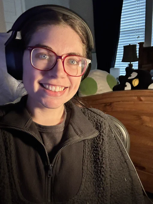 Leah, a woman with brown hair and blue eyes, is wearing headphones and smiling towards the camera. Behind her is a bed with stuffed animals on it.