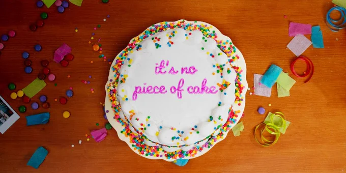 Top-down view of a birthday cake on a table. Icing spells out 'it's no piece of cake'