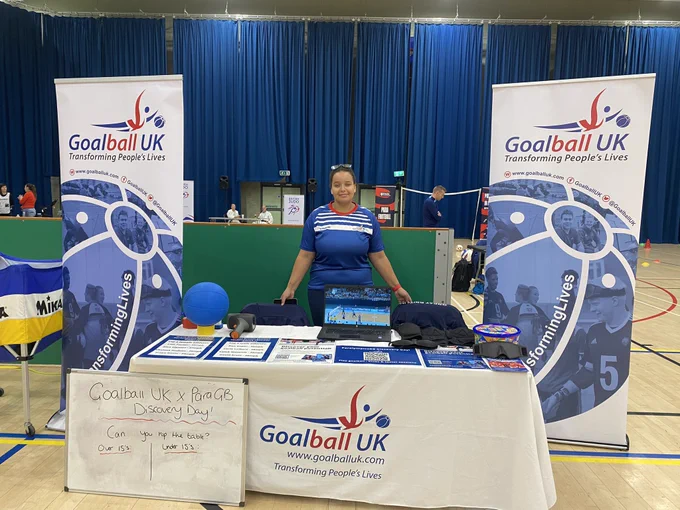 Alex Ulysses standing behind a table at our stand at the Paralympics GB Discovery Day at the K2 Leisure Centre in Crawley. Either side of Alex is 2 Goalball UK banners, and on the table are different leaflets and promotional material.