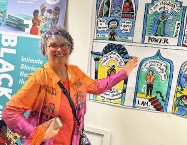 Older silver haired person wearing a bright orange and pink shirt and a multi coloured mohair hat standing next to an panelled illustration which includes a painting of them captioned: “I am stepping into who I fully am”