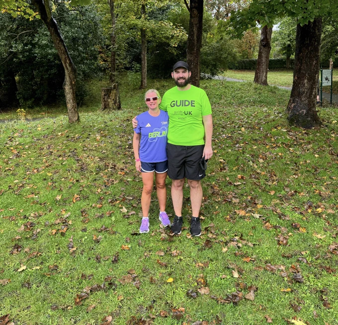 The image shows two people standing on a grassy area with scattered fallen leaves. They are both smiling and appear to be in a park-like setting with trees in the background. The person on the left is a woman wearing sunglasses, a purple T-shirt with the word "BERLIN" in green letters, black shorts, and purple running shoes. The person on the right is a man wearing a bright green T-shirt with the word "GUIDE" in large letters, along with "Running UK" and other text below it, black shorts, and black running shoes. He is also wearing a black cap. The man has his arm around the woman's shoulder.