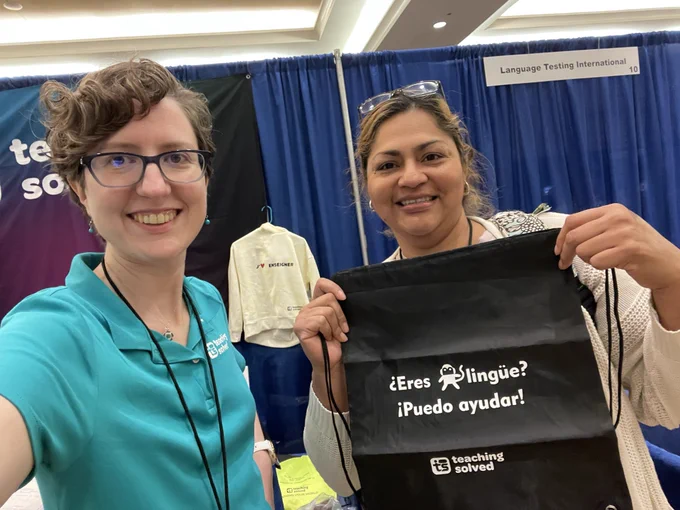 Hope and a teacher holding up a Teaching Solved bag reading “¿Eres monolingüe? Puedo ayudar” in front of the teaching solved booth.