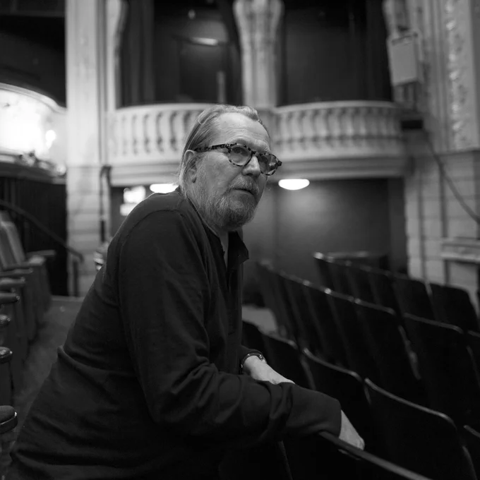 A black and white image of a bearded man in glasses and dark clothes sat in a theatre auditorium