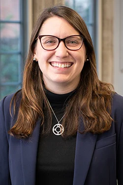 Portrait of a smiling individual, the speaker of the event: Lauren Tilton, wearing glasses, a dark blazer, and a necklace, standing indoors with a window background.