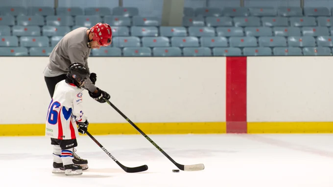 A hockey player wearing a jersey and a helmet practices on an indoor ice rink, with their coach, handling a puck with a stick.